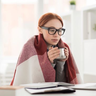 Young woman wearing glasses, wrapped in a blanket holding a cup of a hot drink sitting in front of b