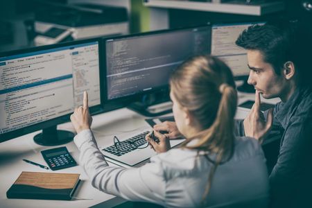 Man and woman programming using three computer monitors