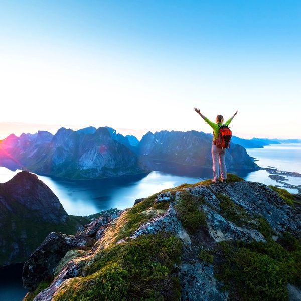 woman standing on mountain top overlooking water and arms overhead in joy as sun rises