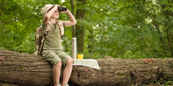 A little boy looking through binoculars and sitting on a log.