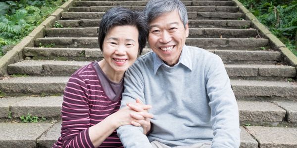 A smiling elderly couple sitting on stairs outside, with woman's hands folded around man's arm