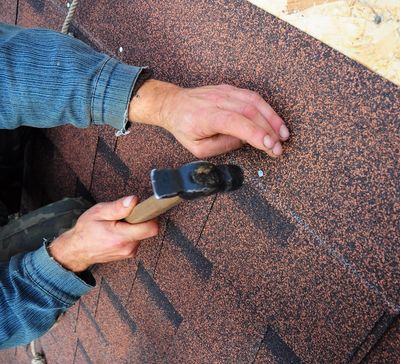 A roofer installing roofing nails into roofing shingles by hand.