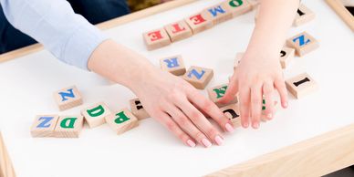 Hands of adult woman on communication therapy board. She forms words with small wood letter blocks 