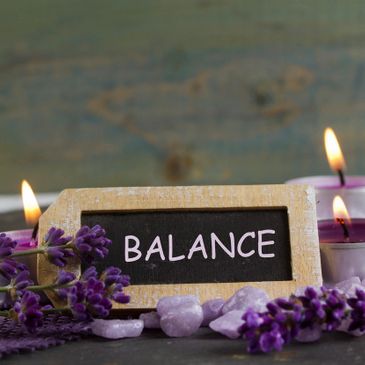 Chaulk board with the word Balance on it with candles along the side of it and purple flowers.