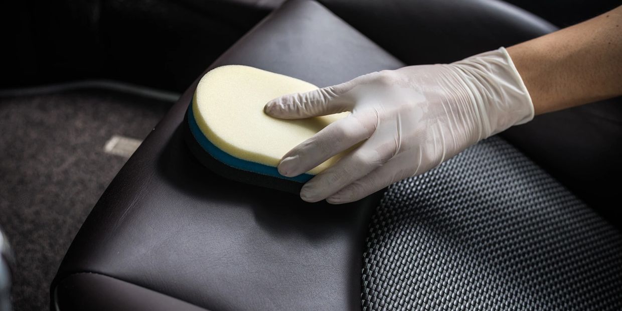 A gloved hand is using a brush to remove dirt from a leather car seat.