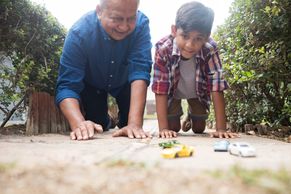 An older gentleman and young boy kneeling on the ground playing with cars. 