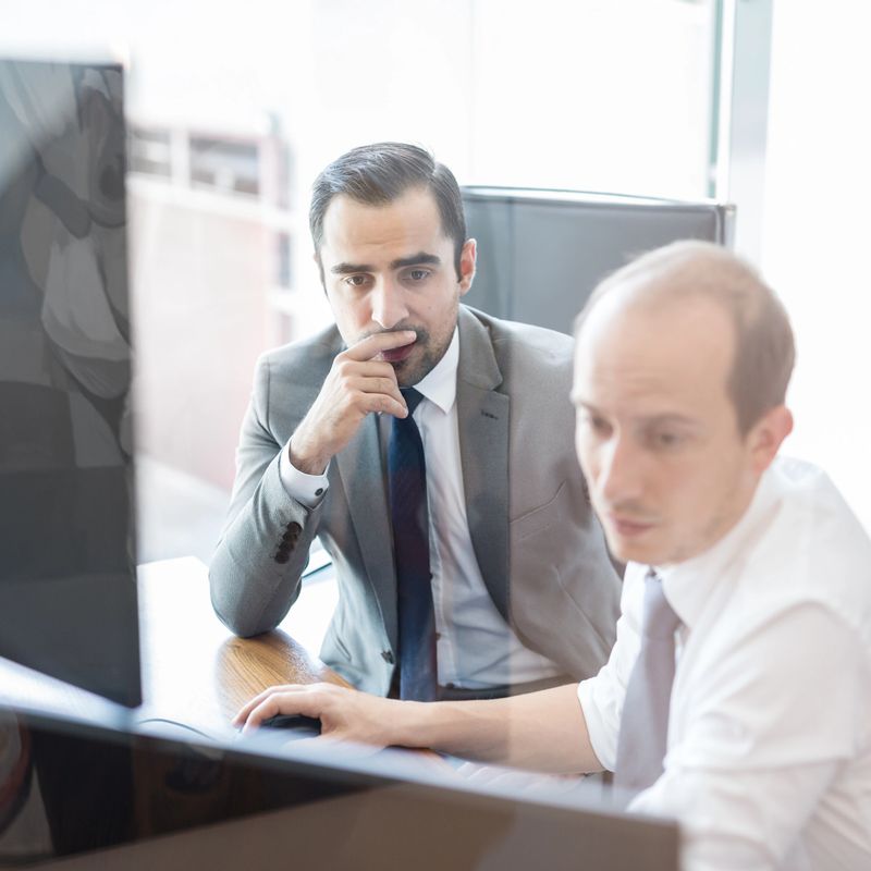 Image of two thoughtful businessmen looking at data on computer screens, solving business issue at business meeting in moder office.