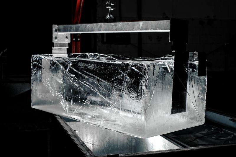 Steel tool holding a huge transparent ice cube on the dark background of modern factory
