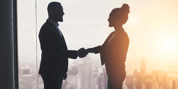 A businessman and businesswoman shaking hands near a large window with a city view during sunrise.