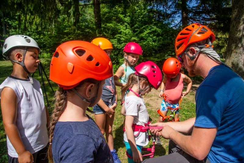A climbing instructor preparing children for a rock climbing camp outside in the summer.  He is attaching a climbing harness to a little girl while he explains safety procedures.
