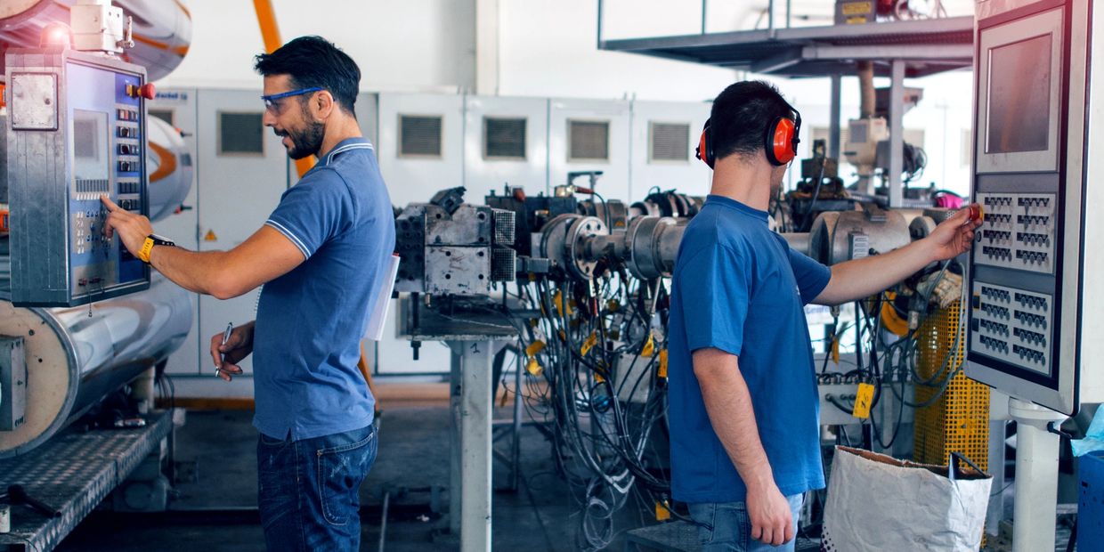 Two factory workers operating machinery control panels in an industrial setting.