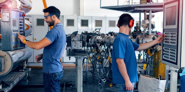 Two factory workers operate machinery in an industrial setting, wearing safety gear.