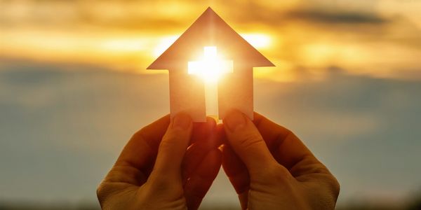 Hands hold a house cutout with a cross-shaped light shining through at sunset.
