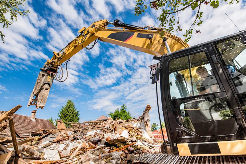 Side view of a yellow excavator in front of a heap of rubble at a demolished building site.