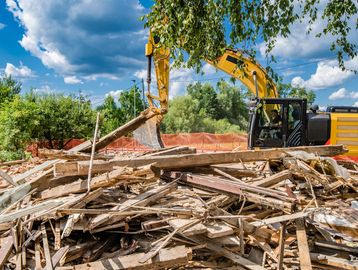 Yellow excavator clearing wooden debris at demolition site under blue sky.