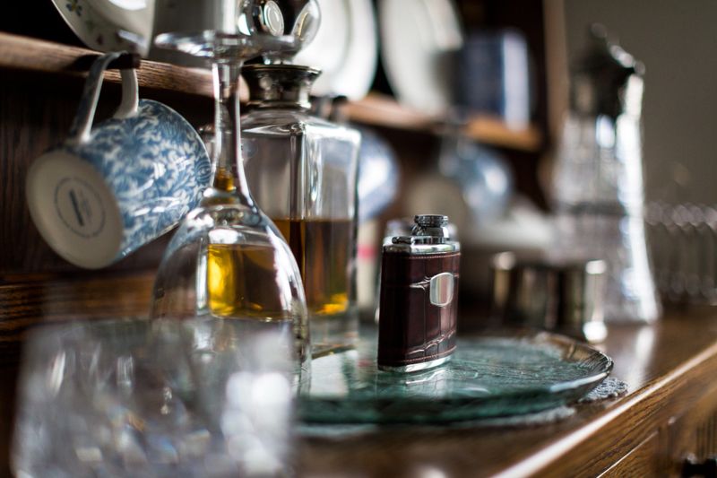Close up color image depicting an arrangement of fine bone china cups and plates, crystal wine and whisky decanters and a leather-bound hip flask on display on an antique oak dresser. Focus is on the hip flask in the center of the image, with the rest of the objects slightly defocused. Room for copy space.