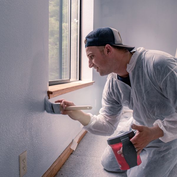 Man in protective suit painting a wall near a window inside a room.