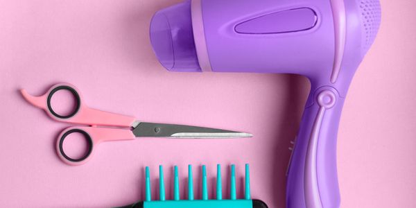 Purple hairdryer, pink scissors, and turquoise comb on pink background.