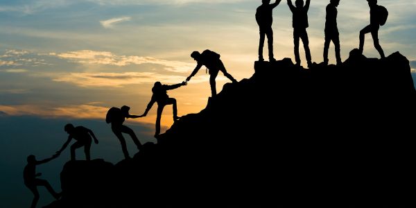 Silhouettes of people helping each other climb a mountain at sunset.