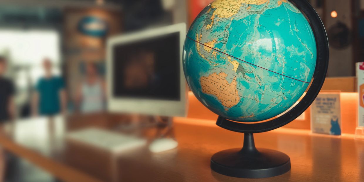 A globe on a desk with a computer and blurred people in the background.