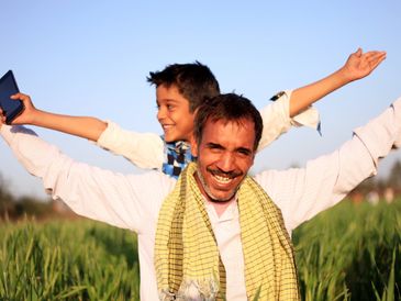 A joyful man carries a smiling boy on his back in a green field under a clear sky.