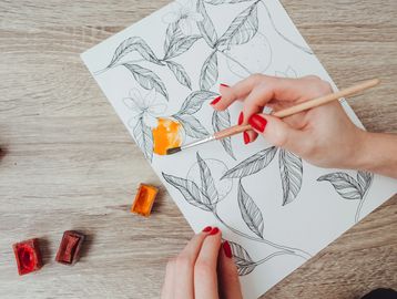 Person painting an orange on a floral sketch with watercolor.