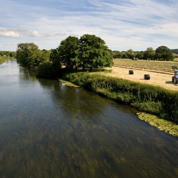 A tranquil river flows beside a farm field with a tractor and hay bales under a blue sky.