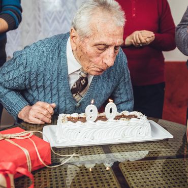 Elderly man blowing out candles on a 90th birthday cake surrounded by family.