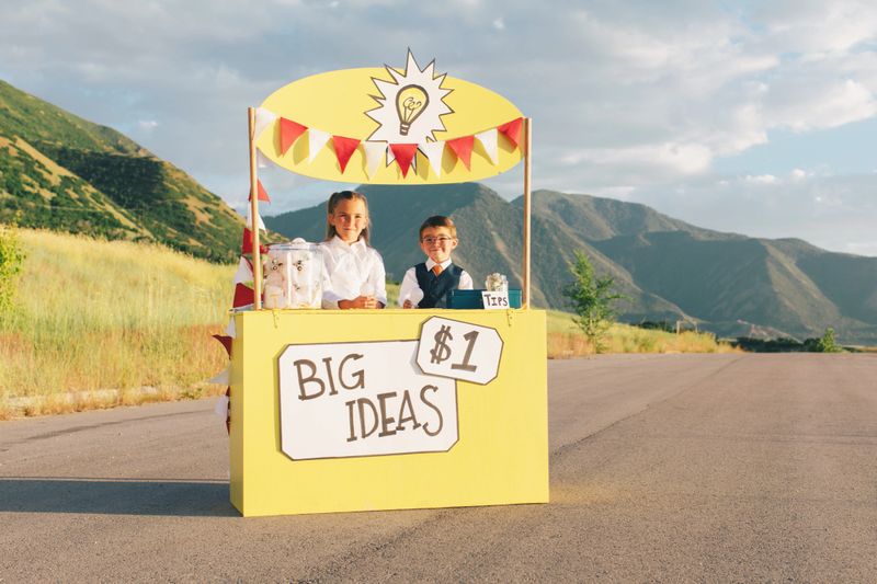Two young children dressed in business attire sit at a lemonade stand turned into a big business idea stand. They are selling the next big and greatest idea and they have lots of them in the form of light bulbs. They have smiles on their faces and are excited to make lots of money for their new business. Image taken in Utah, USA.