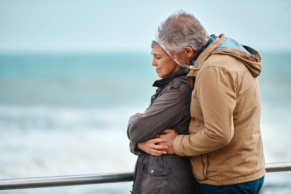 A couple standing looking out to sea on a winter's day, holding each other in a comforting embrace.
