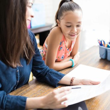 A woman helps a girl with reading or homework at a wooden table.