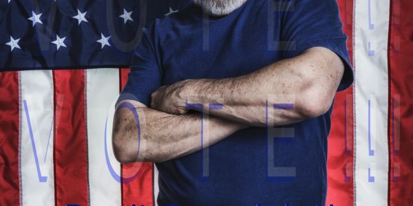 Man with crossed arms stands confidently in front of an American flag with 'VOTE' text overlay.