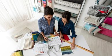 Two women discussing fabric samples and design sketches in a bright studio.