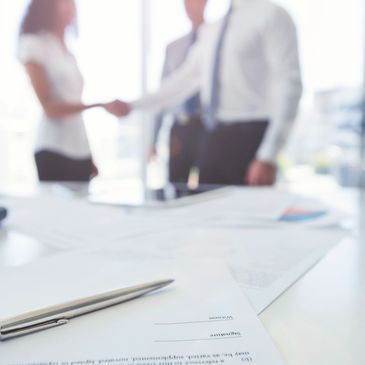 Pen and contract on a desk with businesspeople shaking hands in background.