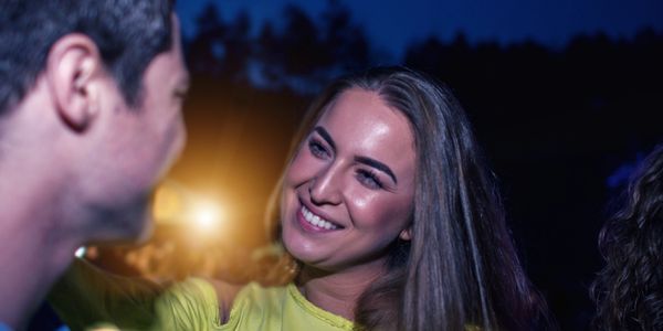 A smiling woman in yellow talks to a man at a nighttime outdoor event with colorful lights.
