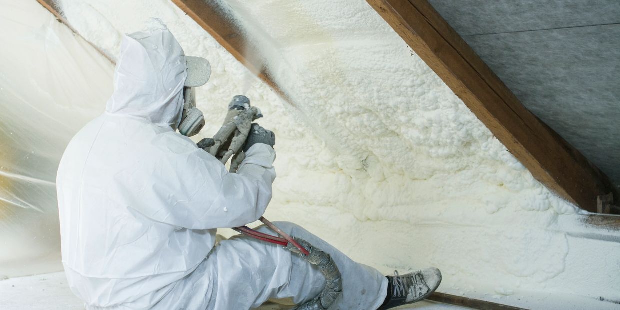 Worker sprays foam insulation in attic wearing protective gear.