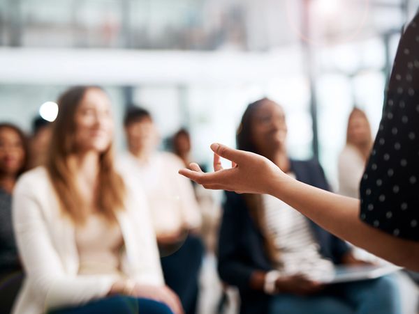 A speaker gestures while addressing an attentive audience in a bright room.