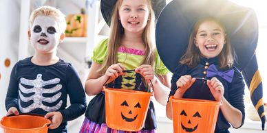 Three children in Halloween costumes holding pumpkin buckets indoors.