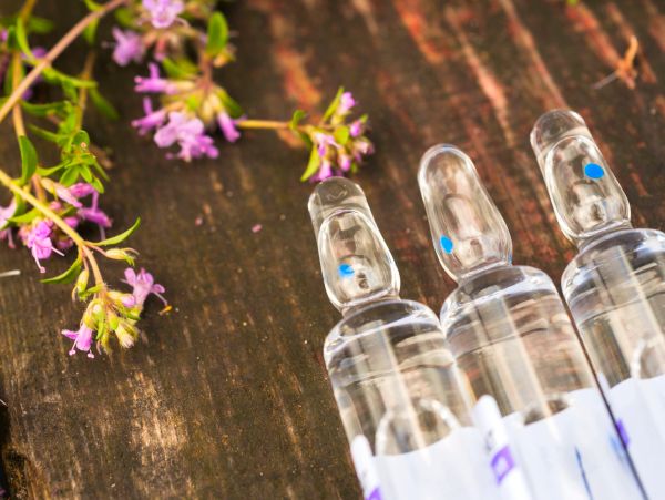 Three glass ampoules with liquid and small purple flowers on wooden surface.