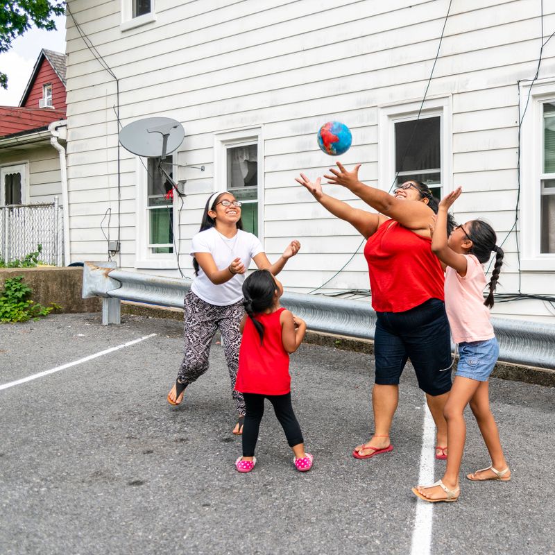 The big happy Latino, Mexican-American family - the mother, body-positive cheerful woman, and kids, girls of different ages - playing with a ball outdoor in the sunny hot summer day at the parking lot nearby his house in Pennsylvania, USA