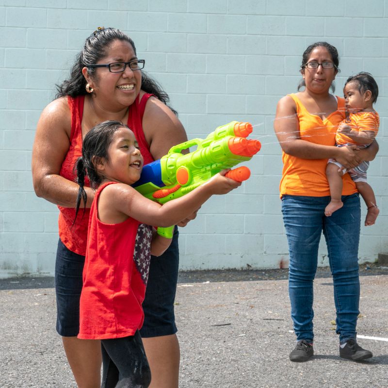 The big happy Latino, Mexican-American family - the mother, body-positive optimistic smiley woman, her sister, and kids, girls of different ages - playing outdoor with a water gun and having fun in the sunny hot summer day at the parking lot nearby his house in Pennsylvania, USA