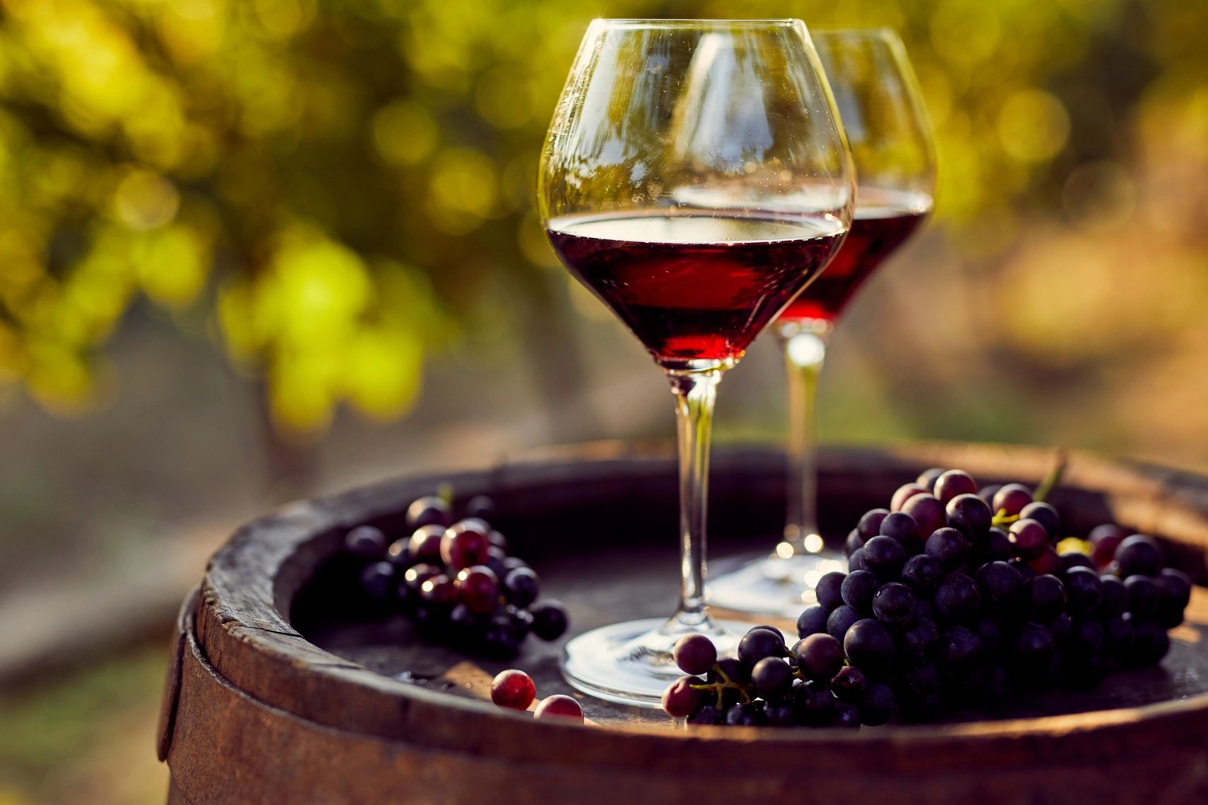 Two glasses of red wine with grapes on a barrel outdoors.