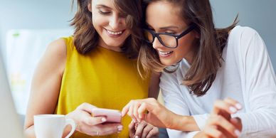 Two women happily looking at a smartphone together at a desk.