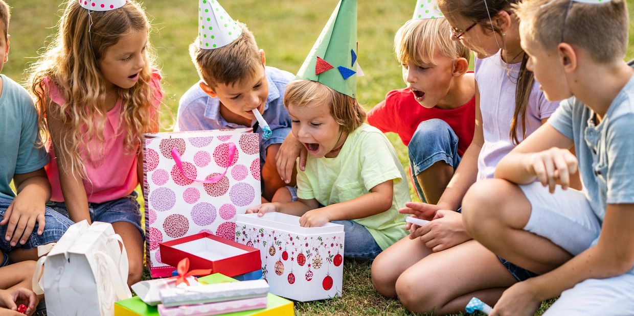 Children wearing party hats excitedly opening gifts at an outdoor birthday party.