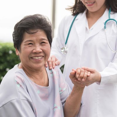 A smiling elderly woman holds hands with a caring doctor outdoors.