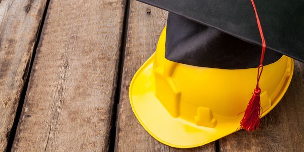 A yellow construction helmet with a black graduation cap on top, placed on wooden surface.