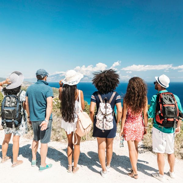 Group of travelers standing together with backpacks and sun hats, enjoying a panoramic coastal view on a group vacation