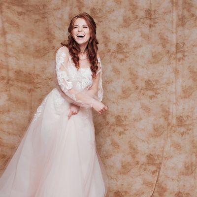 A joyful bride in a lace wedding dress laughs against a textured beige backdrop.