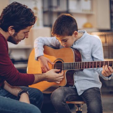 A man teaching a young boy to play acoustic guitar indoors.