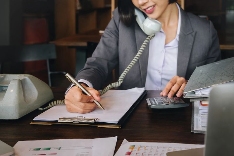 Economist and accountant, female working with document, computer laptop, calculator and using phone contact to customer in office.